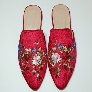 May include: A pair of red velvet mules with floral embroidery. The pointed-toe slip-on shoes feature a cream-colored insole and are decorated with embroidered flowers in white, blue, and red. The shoes are set against a white background.