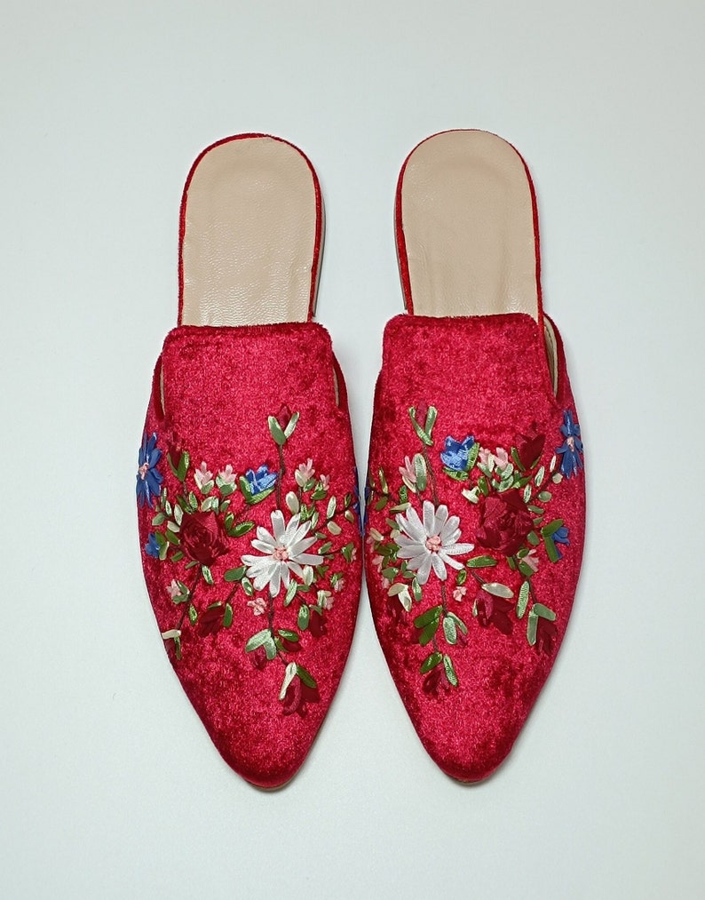 May include: A pair of red velvet mules with floral embroidery. The pointed-toe slip-on shoes feature a cream-colored insole and are decorated with embroidered flowers in white, blue, and red. The shoes are set against a white background.