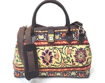 May include: A structured handbag featuring a woven tapestry fabric with a cream, red, blue, and green floral pattern. It includes dark brown leather handles, a matching adjustable shoulder strap, and a gold-tone metal push-lock closure.