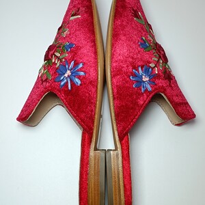 May include: Pair of red velvet slippers with floral embroidery. The slippers have a pointed toe and a low wooden heel. The embroidery features blue, green, and red flowers. The slippers are shown on a white background.