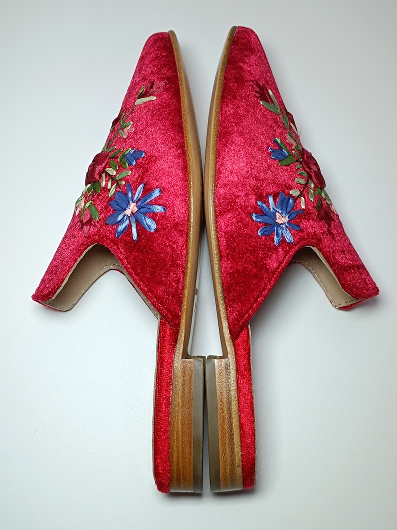 May include: Pair of red velvet slippers with floral embroidery. The slippers have a pointed toe and a low wooden heel. The embroidery features blue, green, and red flowers. The slippers are shown on a white background.