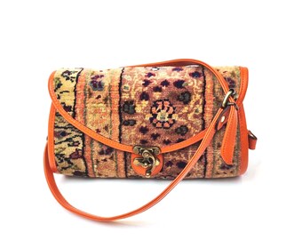 May include: A rectangular shoulder bag featuring a woven fabric body with a multi-colored floral and geometric pattern in shades of orange, brown, and cream. The bag has bright orange leather trim, a matching strap, and a brass metal clasp.