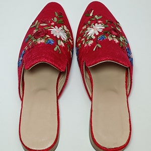 May include: A pair of red velvet slip-on shoes with floral embroidery. The shoes have a pointed toe and a light brown insole. The floral embroidery includes white, blue, and green accents.