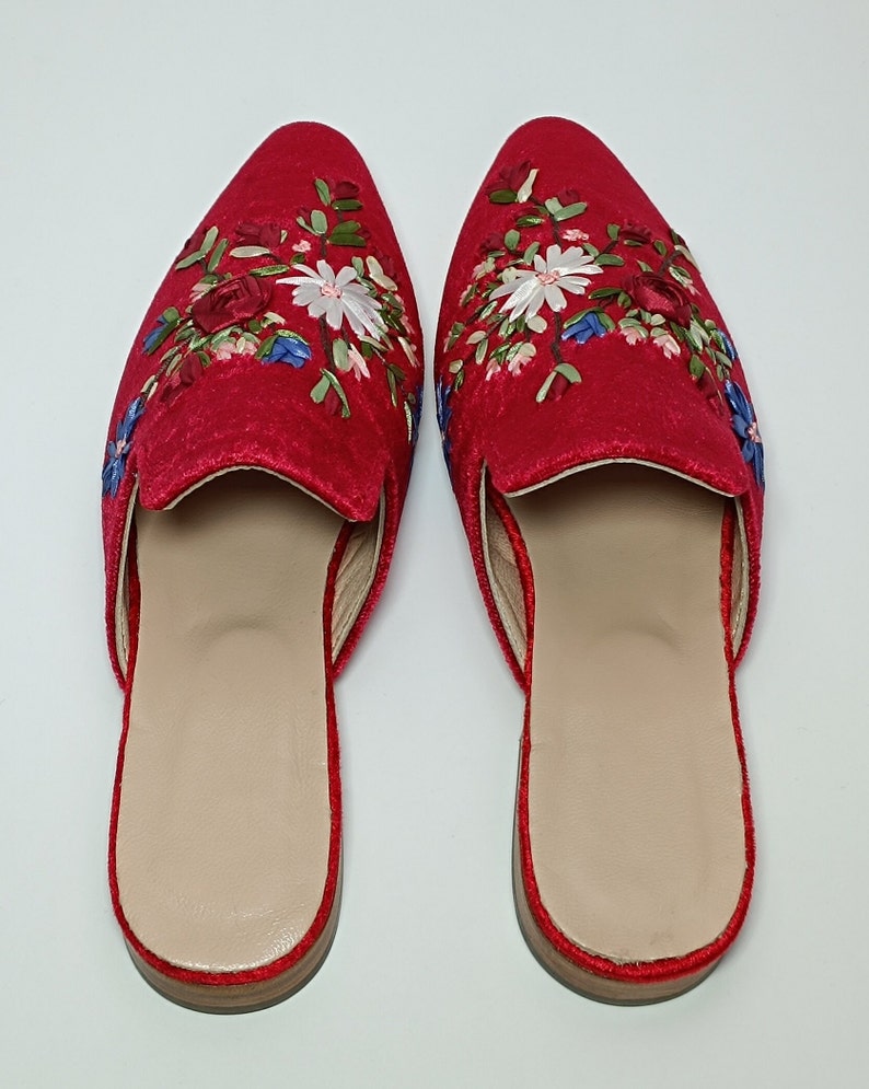 May include: A pair of red velvet slip-on shoes with floral embroidery. The shoes have a pointed toe and a light brown insole. The floral embroidery includes white, blue, and green accents.