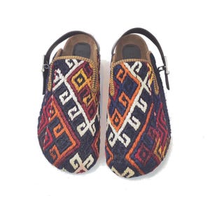 May include: A pair of slip-on shoes featuring a woven textile upper with a geometric pattern in navy blue, red, orange, and white. The shoes include a dark brown leather heel strap with a silver-toned metal buckle and a brown interior footbed.