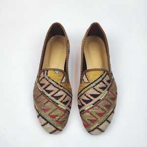 Puede incluir: Un par de zapatos sin cordones con un patrón geométrico colorido. Los zapatos presentan una base beige con un diseño detallado en tonos de amarillo, rojo, verde y marrón. El ribete y el forro interior son de color marrón oscuro.