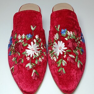 May include: A pair of red velvet slippers with floral embroidery. The embroidery features white daisies, red roses, blue and pink flowers, and green leaves. The slippers have a pointed toe and a low heel.