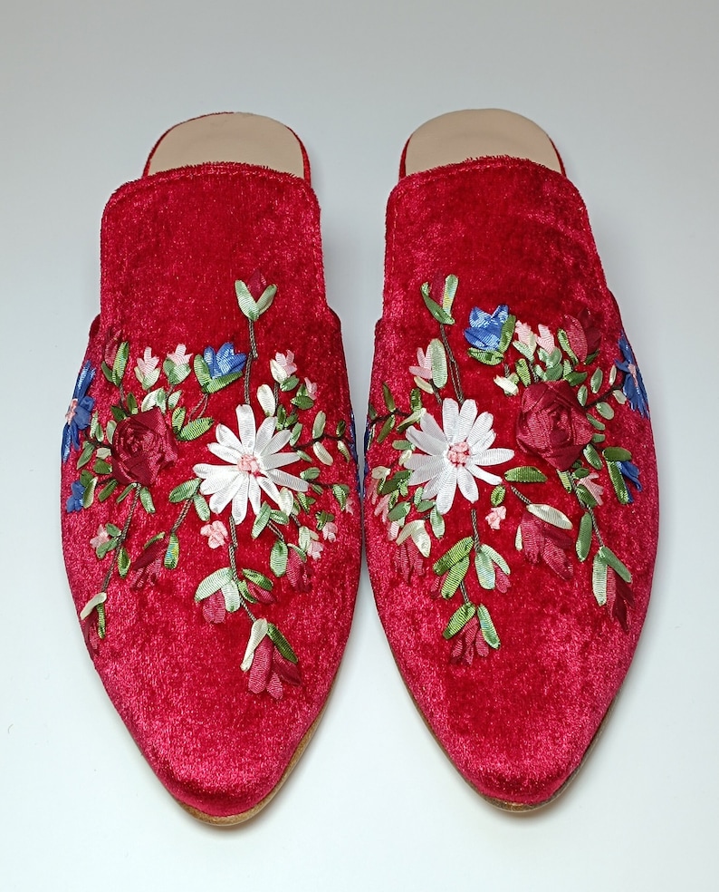 May include: A pair of red velvet slippers with floral embroidery. The embroidery features white daisies, red roses, blue and pink flowers, and green leaves. The slippers have a pointed toe and a low heel.