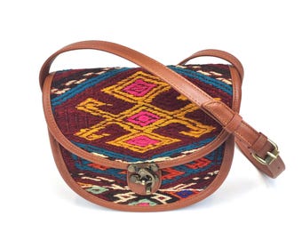 May include: A saddle-shaped crossbody bag featuring a woven textile flap with a geometric pattern in shades of deep red, bright pink, yellow, and teal. The bag is trimmed with brown leather piping and includes a matching adjustable leather strap and a brass-toned metal clasp closure.