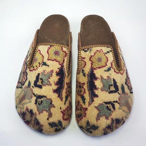 Op de afbeelding: Een paar beige pantoffels met een bloemenpatroon. De pantoffels hebben een bruine leren zool en een gouden rand rond de opening.