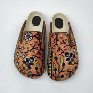 May include: A pair of slip-on mules featuring a textured, peach-colored fabric upper with a detailed floral pattern in navy blue, green, and yellow. The shoes have a cream-colored interior lining, a dark brown heel patch, and brown trim.