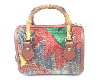 May include: A small handbag featuring a woven textile body with a multi-colored geometric pattern in red, green, yellow, and blue. The bag includes brown leather trim, gold-tone metal hardware, and a curved bamboo handle attached with leather tabs.