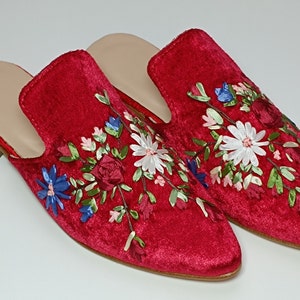 May include: A pair of red velvet slip-on shoes with floral embroidery. The shoes feature a light-colored insole and a low heel. The floral design includes white daisies, blue and pink accents, and green leaves.