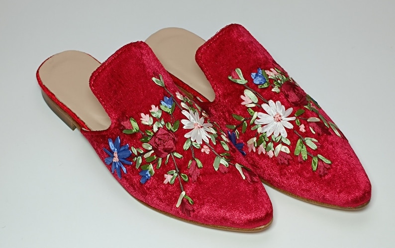 May include: A pair of red velvet slip-on shoes with floral embroidery. The shoes feature a light-colored insole and a low heel. The floral design includes white daisies, blue and pink accents, and green leaves.