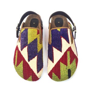 May include: A pair of slip-on shoes featuring a woven textile upper with a geometric pattern in red, green, purple, and cream. The shoes include a brown leather heel strap with a metal buckle and a black insole marked with the number 97.