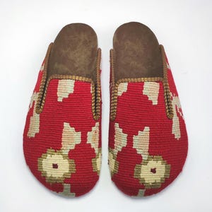 Op de afbeelding: Een paar rode en witte gepatroneerde pantoffels met bruine suède inlegzolen. De pantoffels hebben een gouden rand rond de randen.