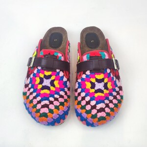 May include: A pair of colorful crochet clogs. The clogs feature a vibrant patchwork design in shades of pink, blue, yellow, and green. They have brown leather straps with silver buckles and a wooden footbed. The clogs are size 41.