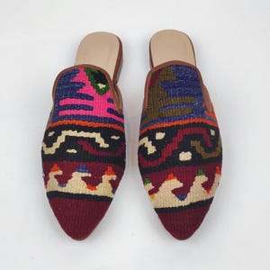 May include: A pair of colorful, patterned slip-on shoes. The shoes feature a pointed toe and a woven design with shades of pink, blue, and burgundy. The interior is a light tan color, and the trim is brown.
