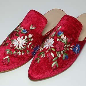May include: A pair of red velvet slip-on shoes with floral embroidery. The embroidery features white, blue, and pink flowers with green leaves. The shoes have a light-colored insole and a flat sole.
