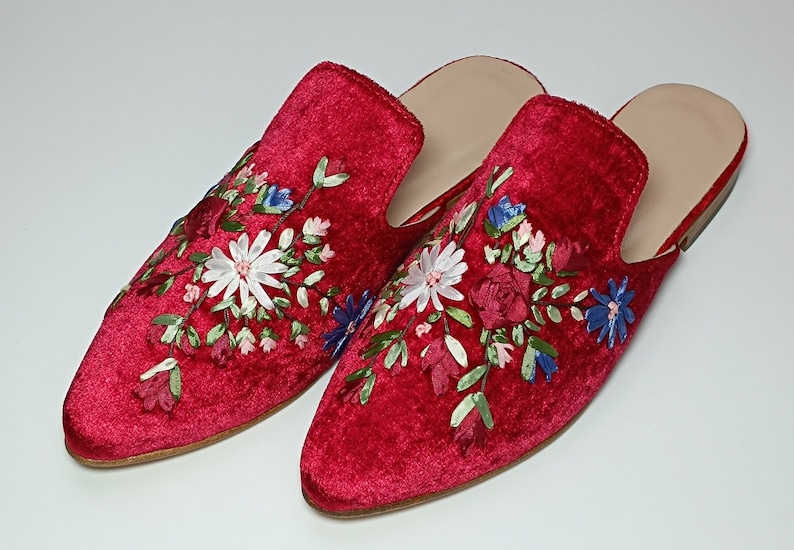 May include: A pair of red velvet slip-on shoes with floral embroidery. The embroidery features white, blue, and pink flowers with green leaves. The shoes have a light-colored insole and a flat sole.