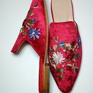 May include: A pair of red velvet mules with floral embroidery. The shoes have a pointed toe and a small block heel. The embroidery features blue, white, and pink flowers with green leaves. The interior of the shoe is a light beige color.