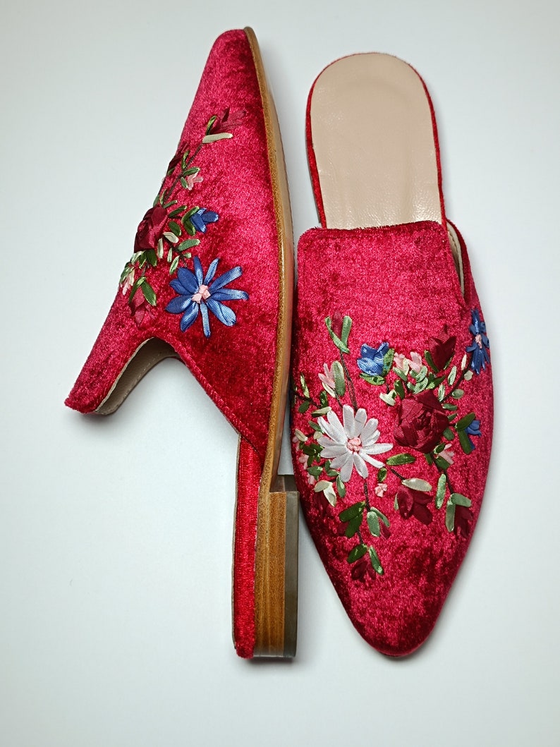May include: A pair of red velvet mules with floral embroidery. The shoes have a pointed toe and a small block heel. The embroidery features blue, white, and pink flowers with green leaves. The interior of the shoe is a light beige color.