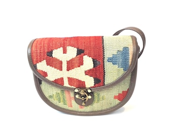 May include: A saddle-shaped crossbody bag crafted from woven kilim fabric featuring a geometric pattern in red, cream, green, and blue. The bag is trimmed with brown leather piping and includes a gold-toned metal latch closure on the front flap.
