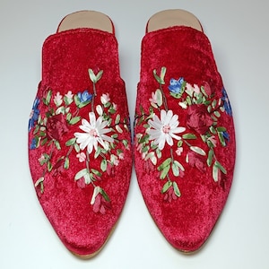 May include: A pair of red velvet mules with floral embroidery. The embroidery features white, blue, and burgundy flowers with green leaves. The shoes have a pointed toe and a low heel.