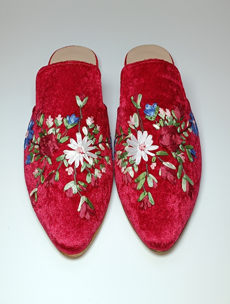 May include: A pair of red velvet mules with floral embroidery. The embroidery features white, blue, and burgundy flowers with green leaves. The shoes have a pointed toe and a low heel.