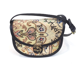 May include: A saddle-shaped crossbody bag featuring a cream-colored tapestry fabric with a multi-colored paisley and floral pattern. The bag is trimmed with dark brown leather piping and an adjustable strap, secured by a brass-toned metal clasp.