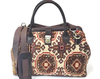 May include: A structured handbag featuring a cream-colored fabric with a repeating geometric pattern in shades of brown, red, and black. It includes dark brown leather handles, a central metal push-lock closure, and a detachable brown fabric shoulder strap.