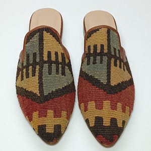 May include: A pair of pointed-toe, flat mules with a geometric pattern in shades of red, brown, beige, and blue. The mules have a brown leather trim around the opening.