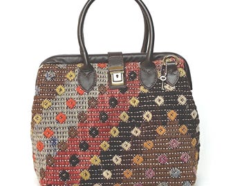 May include: A structured handbag featuring a woven textile body with a multi-colored diamond pattern in shades of red, brown, black, and gray. It includes dark brown leather handles, a central metal clasp closure, and a small metal key attached.