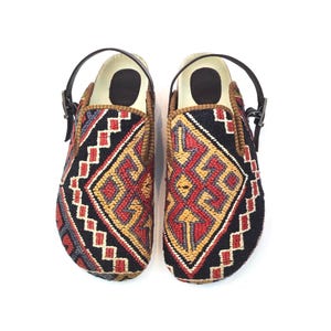 May include: A pair of slip-on shoes featuring a woven textile upper with a geometric pattern in red, yellow, and black. The shoes include a dark brown leather heel strap with a metal buckle and a light-colored interior footbed.