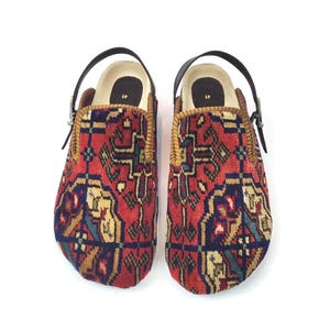 May include: A pair of slip-on shoes featuring a red, blue, and beige woven textile pattern. Each shoe has a black adjustable heel strap with a metal buckle and a black insole printed with the number 41. The edges are finished with a tan trim.