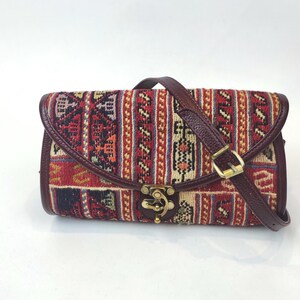 Puede incluir: Bolso rectangular con un diseño de tela estampada en rojo, beige y azul. El bolso tiene un ribete de cuero burdeos, una correa para el hombro a juego y un cierre dorado. El bolso se cierra con una solapa.