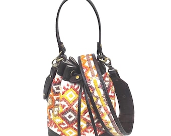Op de afbeelding: Een bucket bag met een geweven stoffen body met een geometrisch patroon in oranje, gele en rode tinten. De tas is voorzien van donkerbruine lederen afwerking, een trekkoordsluiting, een handvat en een afneembare brede schouderriem.
