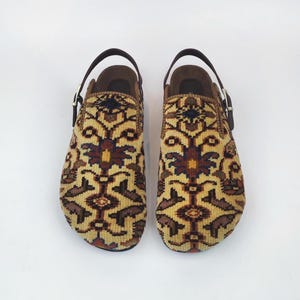 May include: A pair of clogs with a patterned textile design in shades of tan, brown, and navy blue. The clogs have a brown leather strap and buckle. The interior is a brown suede-like material.