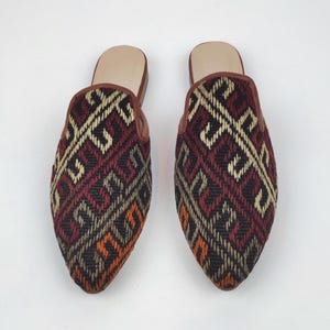 May include: A pair of woven textile slippers with a geometric pattern in burgundy, black, beige, and orange. The slippers have a pointed toe and a brown leather trim and insole.