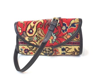 May include: A rectangular shoulder bag featuring a woven tapestry fabric with a red, yellow, and blue floral paisley pattern. The bag has dark brown leather trim, a matching adjustable shoulder strap, and a brass-toned metal clasp closure.