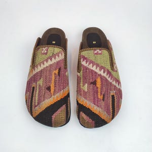 May include: A pair of patterned slip-on shoes. The shoes feature a woven design with shades of purple, green, orange, and black. The interior is brown, and the soles are a darker brown. The shoes are displayed on a white background.