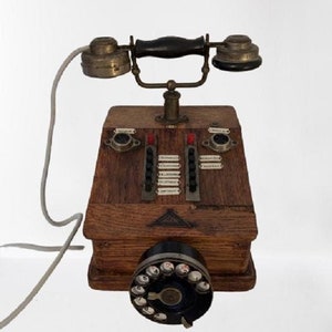 Puede incluir: Un teléfono de disco rotatorio de madera vintage con un auricular negro y detalles en latón. El teléfono tiene una caja de madera con una serie de botones y un disco rotatorio.