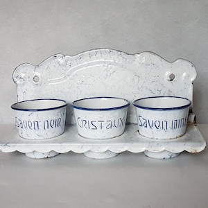 Peut inclure: Porte-savon mural en émail blanc avec trois compartiments. Chaque compartiment a un bord bleu et une inscription bleue. Les compartiments sont étiquetés "Savon Noir", "Cristaux" et "Savon Jaune".