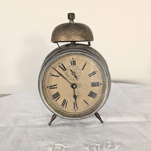 Könnte beinhalten: Antiker Wecker mit ovaler Form, Metallgehäuse und Glocke oben. Das Zifferblatt zeigt römische Ziffern und ein kleineres Zifferblatt. Die Zeiger sind schwarz, und die Uhr steht auf drei kleinen Beinen.
