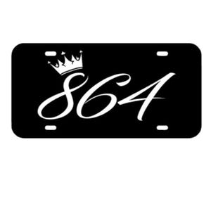 May include: Black license plate with the number 864 in white script, topped with a white crown. The plate has rounded corners and small holes for mounting.