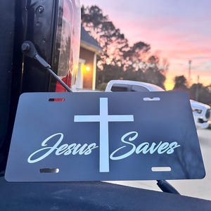 May include: Black license plate with a white cross and the text "Jesus Saves" in a white cursive font. The plate is mounted on a black vehicle.