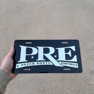 May include: A rectangular black license plate featuring the white text PRE in a large bold font. Below the letters, a white banner displays the text PAPER ROUTE EMPIRE with a small star on each side. The plate has four mounting slots at the corners.