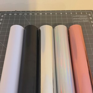 May include: Five rolls of adhesive vinyl arranged side by side. The colours from left to right are matte white, matte black, glossy white, iridescent silver with a rainbow sheen, and a metallic rose gold finish, all resting on a cutting mat.