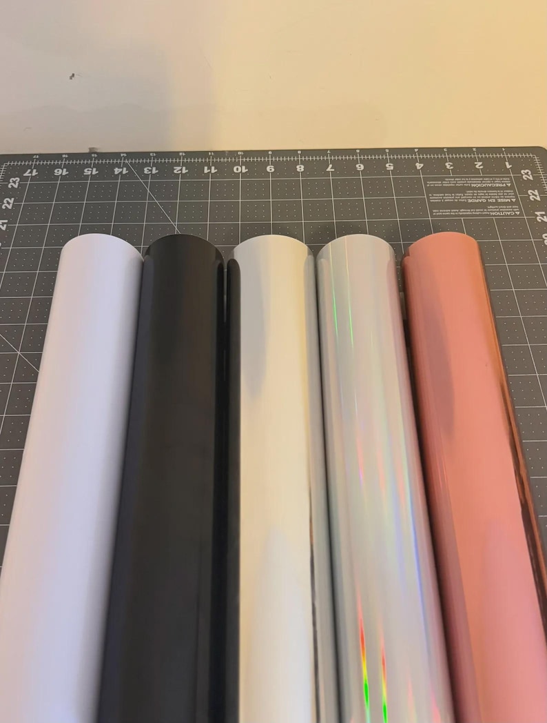 May include: Five rolls of adhesive vinyl arranged side by side. The colours from left to right are matte white, matte black, glossy white, iridescent silver with a rainbow sheen, and a metallic rose gold finish, all resting on a cutting mat.