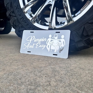 May include: White metal license plate with the text "Pimpin' Ain't Easy" and a graphic of three stick figures. The license plate is sitting on a gray surface.